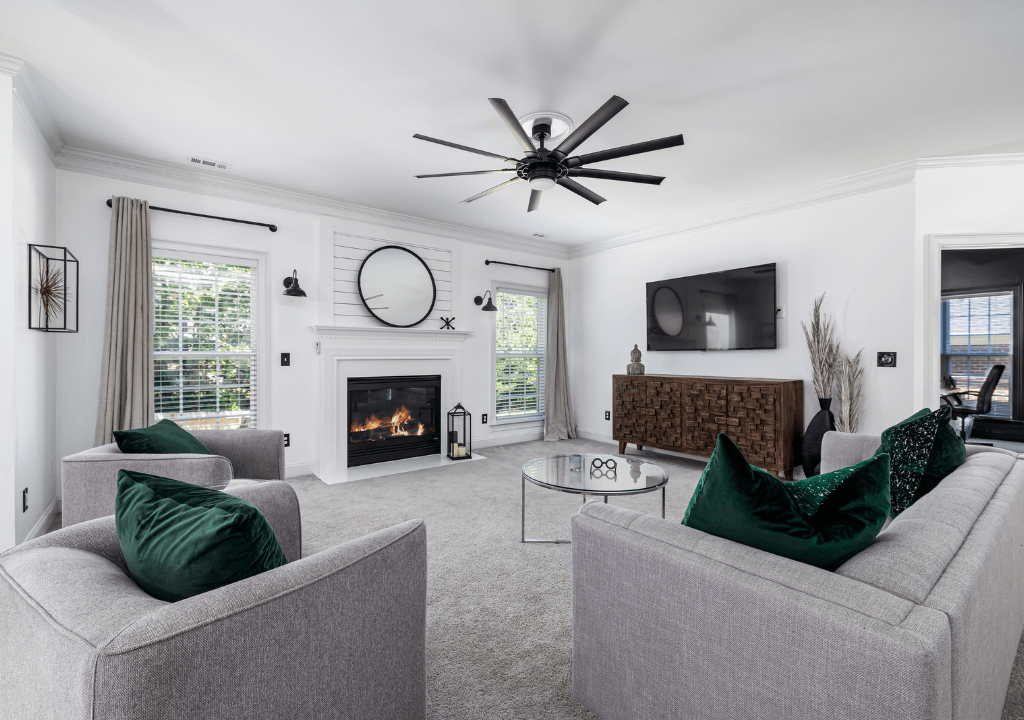 Modern living room with white walls, gray furniture, emerald green velvet pillows, wood console, and a central fireplace.