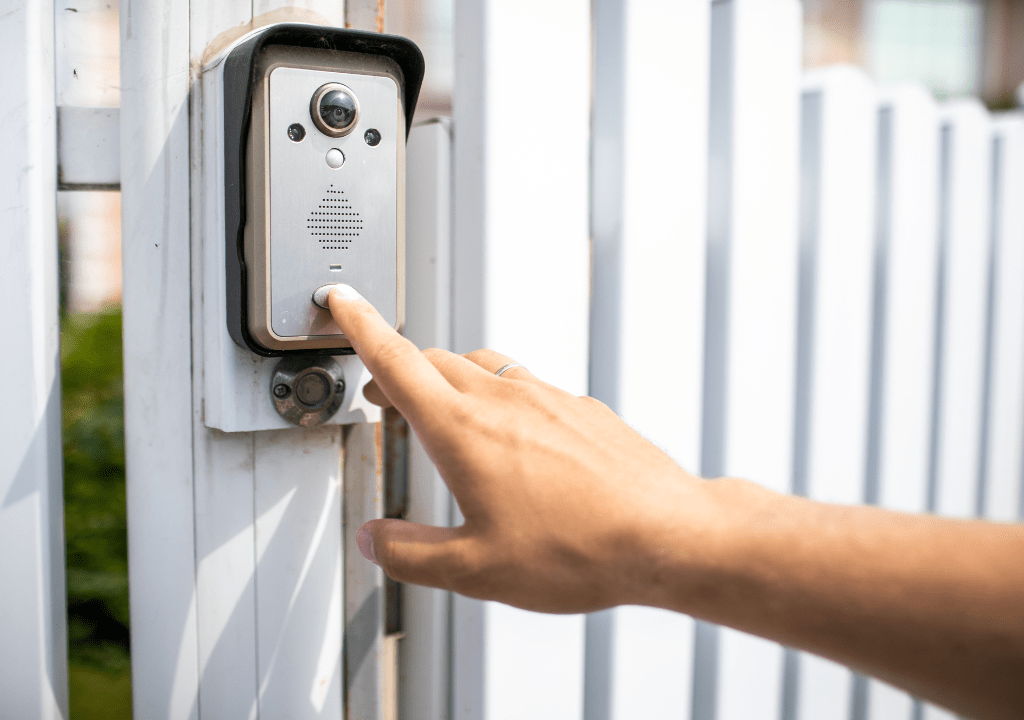 Close-up of a person's hand pressing the button on a silver video intercom or doorbell mounted on a white fence or gate.